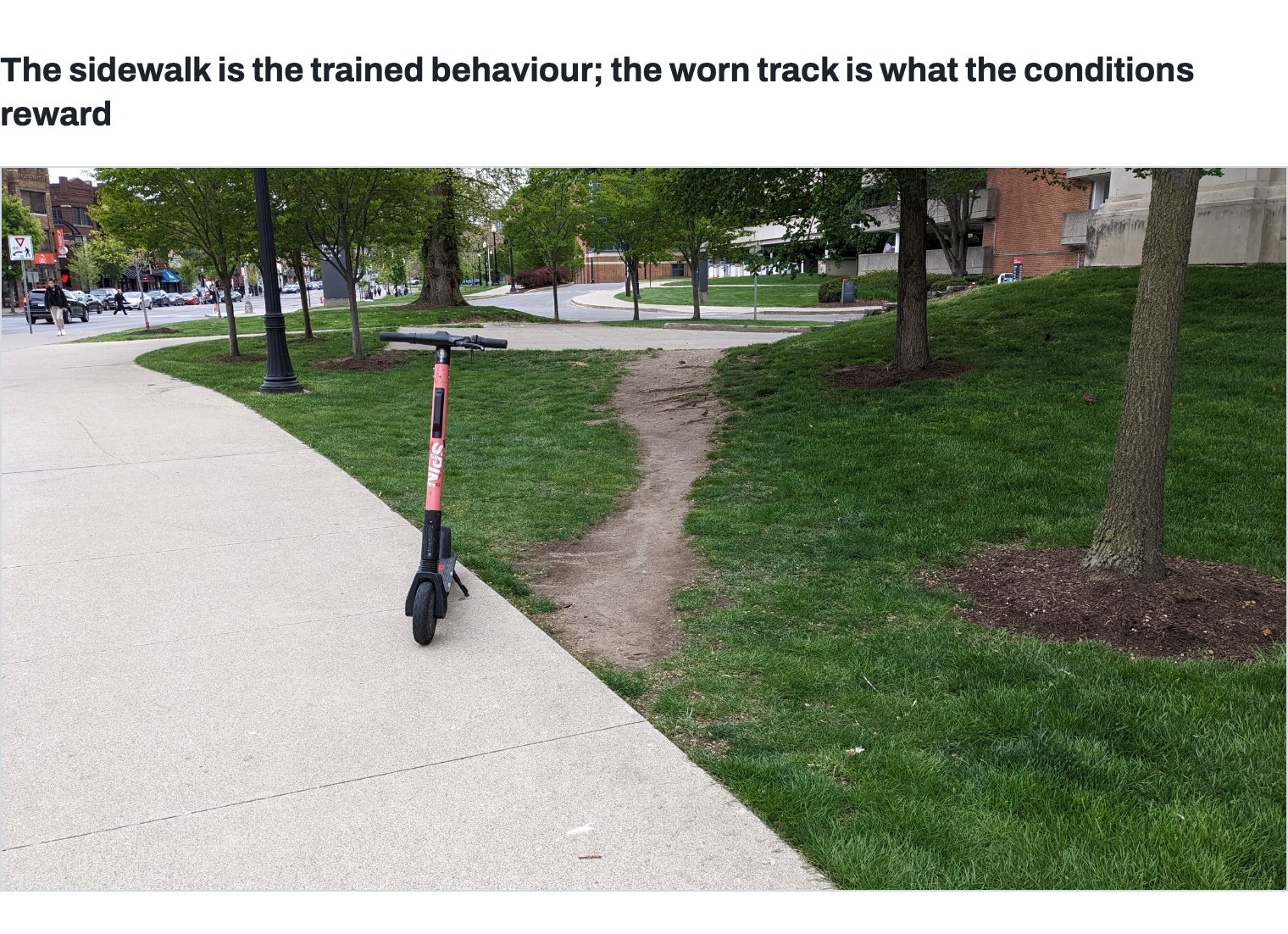 A paved path curving across a lawn. A narrow, winding track of bare earth is worn through the grass beside it, cutting across to the walkway beyond where people actually walk. An e-scooter stands at the edge of the paving.